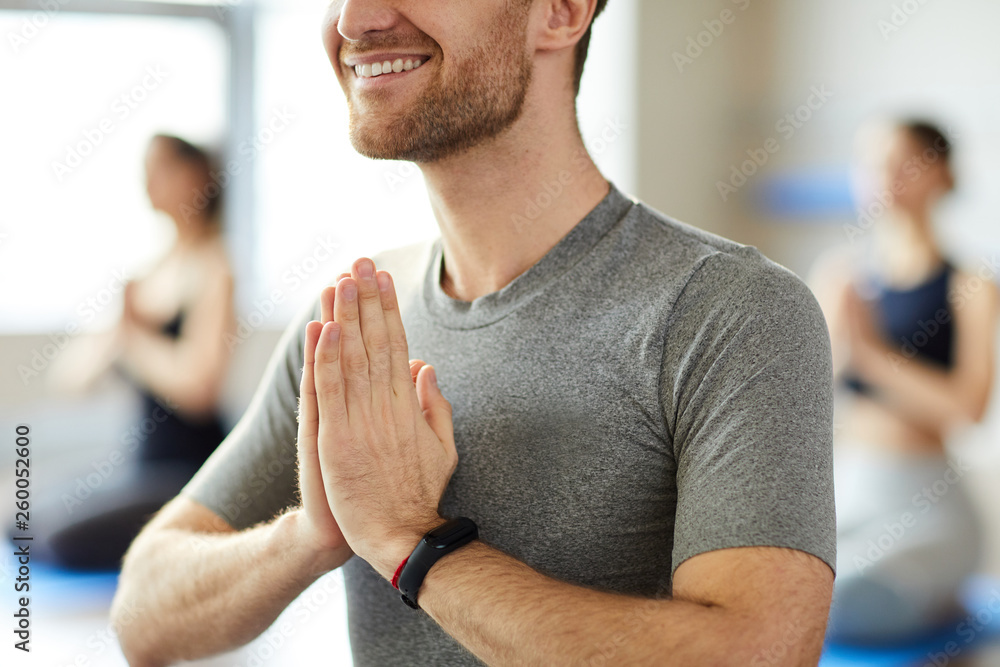 Fototapeta premium Close-up of smiling young man with stubble wearing smartband holding hands in Namaste while greeting yoga teacher at class