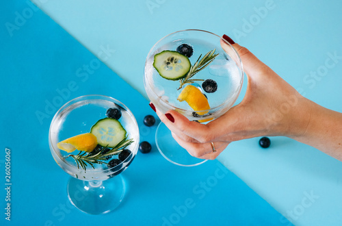 Woman holding a gin and tonic drink with blueberries, cucumber and rosemary on a blue geometric, colorful background.
