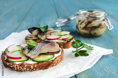 smorrebrod sandwich with herring rye bread sliced cucumber radish parsley on paper salted fish in jar on blue wooden background photo