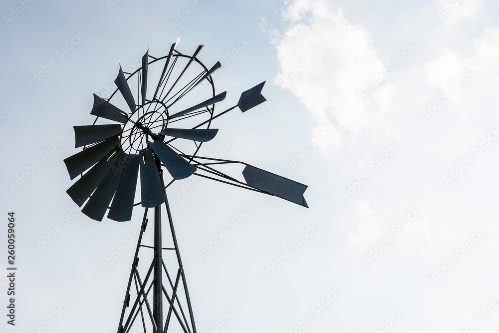 Low angle view of an old-fashioned, multi-bladed, metal wind pump atop a lattice tower in backlight against a pale blue sky with white clouds. Low angle view of an old-fashioned, multi-bladed, metal wind pump atop a lattice tower in backlight against a pale blue sky with white clouds.