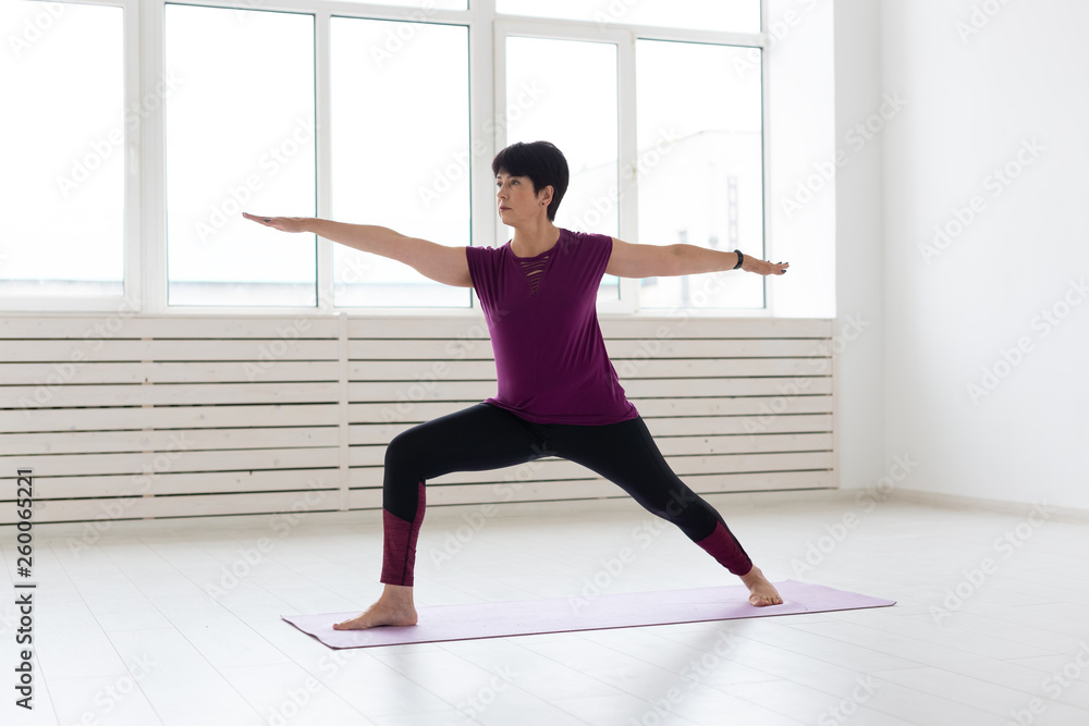Fototapeta premium People, yoga, sport and healthcare concept - Middle-aged woman standing in Warrior over white background