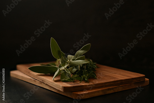 A bunch of greens for cooking meat consisting of rosemary and sage lies on a wooden cutting board. Light hightlights the leaves shine. Dark background. photo