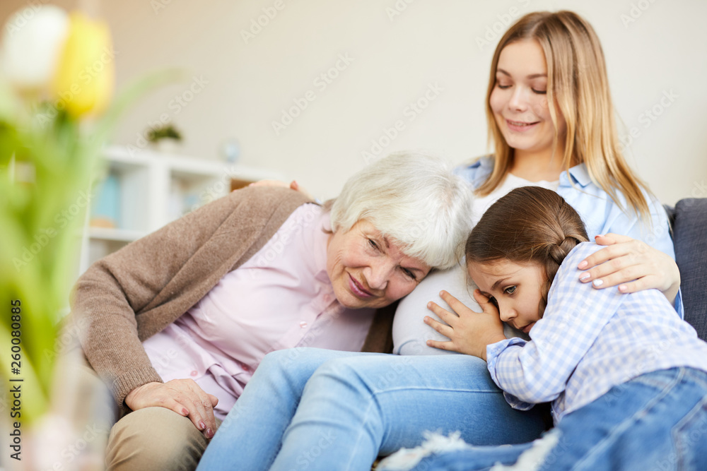 Portrait of grandmother and little girl listening to pregnant belly of smiling young woman, copy space