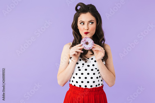 Photo of feminine pin-up woman 20s in vintage polka dot dress smiling while holding and eating tasty donut