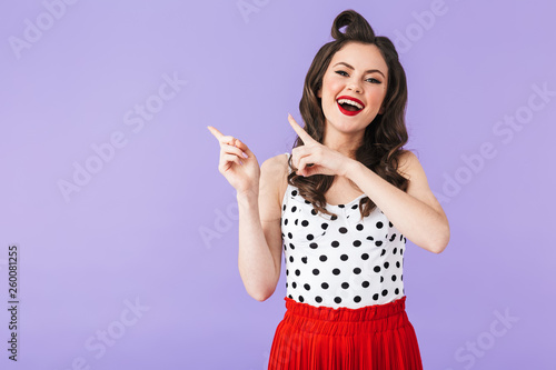 Portrait of cheery pin-up woman 20s in vintage polka dot dress pointing fingers aside at copyspace