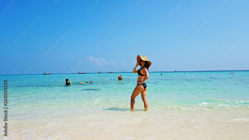 The girl in the hat and swimsuit on the beach. Glasses on. European woman on the beach with white sand. The water is turquoise with a blue tint. Photo shoot model. White sand and blue sky. Paradise.