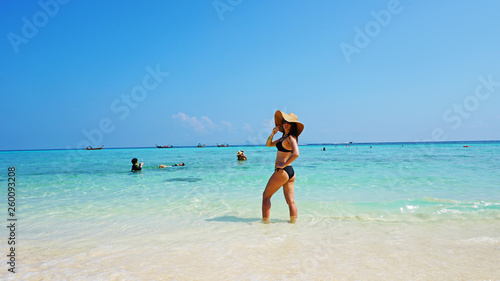The girl in the hat and swimsuit on the beach. Glasses on. European woman on the beach with white sand. The water is turquoise with a blue tint. Photo shoot model. White sand and blue sky. Paradise.
