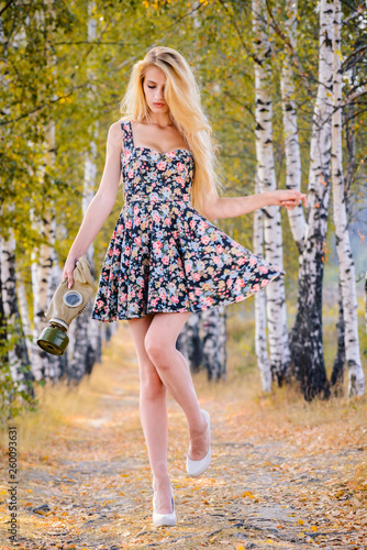 An attractive girl with long hair in a short dress and heels with a gas mask in her hands walks in the Park.