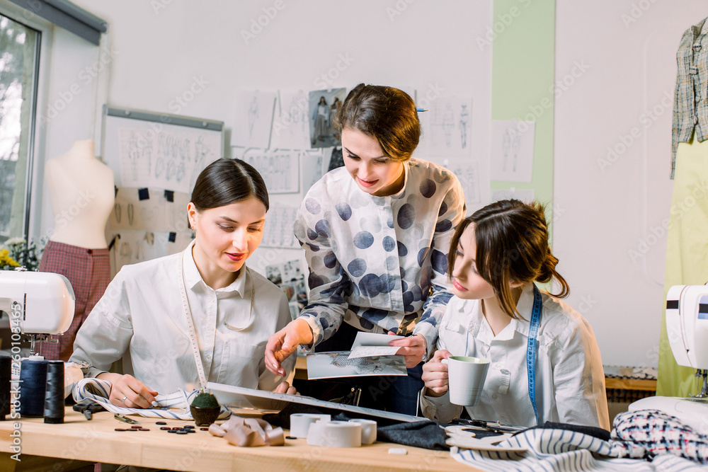 Fashion Design, dressmaker, tailor Concept. Three young Caucasian women seamstresses working together at the bright atelier, preparing new handmade clothes collection