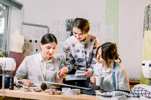 Fashion Design, dressmaker, tailor Concept. Three young Caucasian women seamstresses working together at the bright atelier, preparing new handmade clothes collection