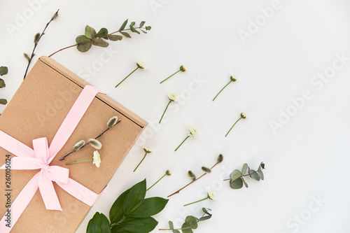 Spring composition with a gift box with a pink bow and green sprigs and flowers lying on a white background. Background, copy space for text, top view, close-up