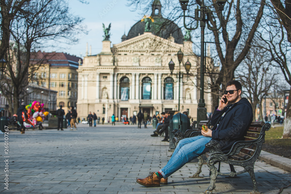 Obraz premium man eating burger surfing internet on phone sitting on bench