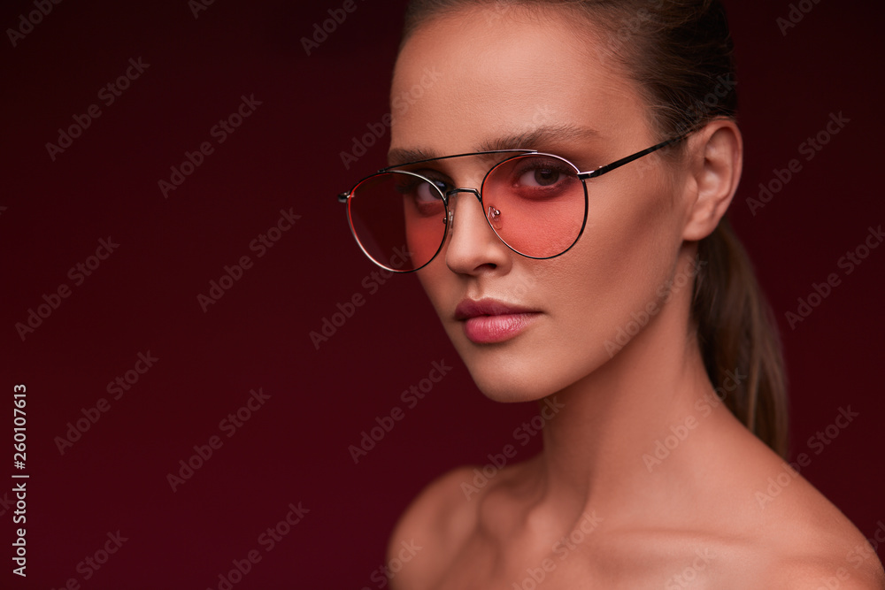 Fototapeta premium Portrait of beautiful young woman with perfect makeup wearing pink sunglasses. Smiling fashion model in aviator sunglasses posing on burgundy background. Studio shot. Summer vacation.