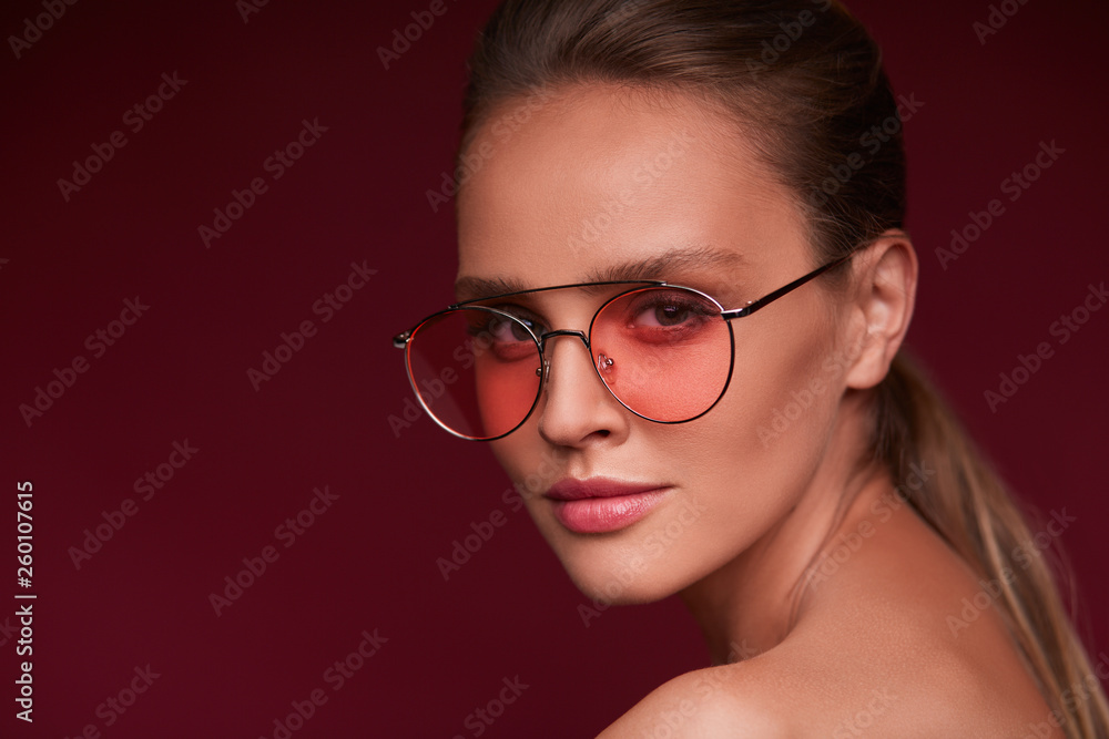 Portrait of beautiful young woman with perfect makeup wearing pink sunglasses. Smiling fashion model in aviator sunglasses posing on burgundy background. Studio shot. Summer vacation. Portrait of beautiful young woman with perfect makeup wearing pink sunglasses. Smiling fashion model in aviator sunglasses posing on burgundy background. Studio shot. Summer vacation.