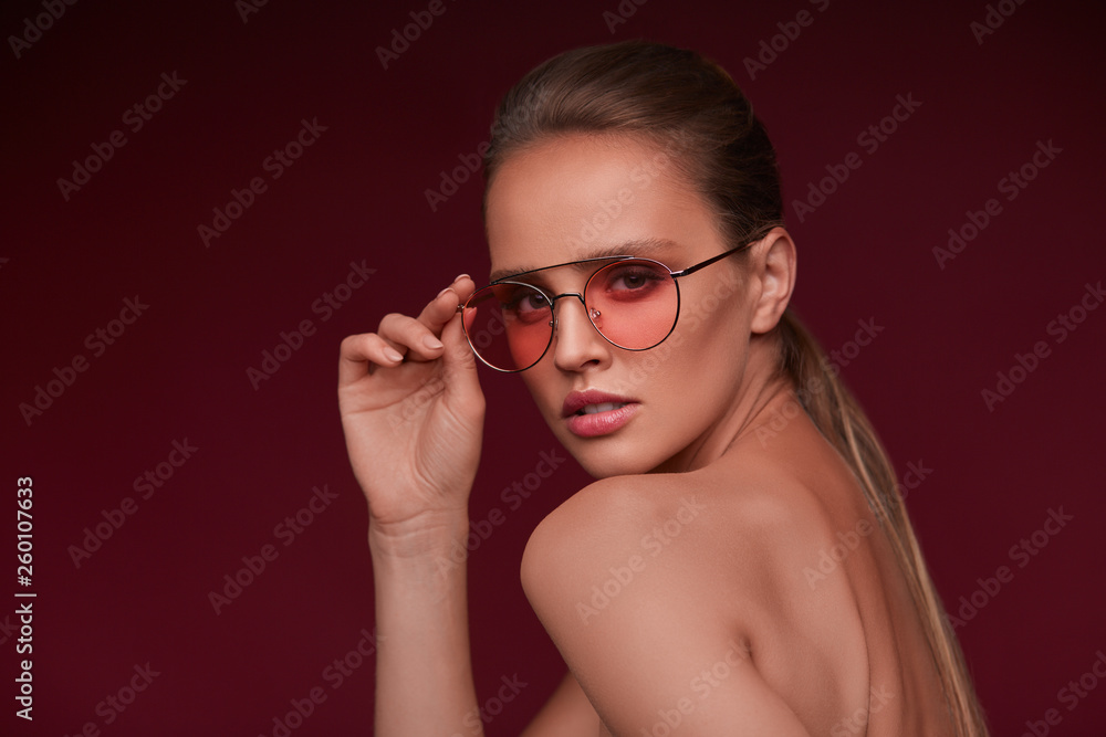 Portrait of beautiful young woman with perfect makeup wearing pink sunglasses. Smiling fashion model in aviator sunglasses posing on burgundy background. Studio shot. Summer vacation. Portrait of beautiful young woman with perfect makeup wearing pink sunglasses. Smiling fashion model in aviator sunglasses posing on burgundy background. Studio shot. Summer vacation.