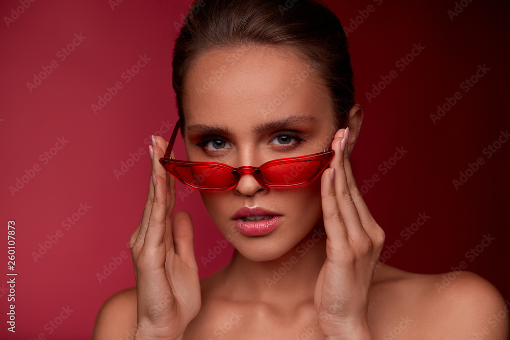 Portrait of beautiful young woman with perfect makeup wearing red sunglasses. Smiling fashion model in cat eye sunglasses posing on burgundy background. Studio shot. Summer vacation. Portrait of beautiful young woman with perfect makeup wearing red sunglasses. Smiling fashion model in cat eye sunglasses posing on burgundy background. Studio shot. Summer vacation.