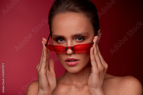 Portrait of beautiful young woman with perfect makeup wearing red sunglasses. Smiling fashion model in cat eye sunglasses posing on burgundy background. Studio shot. Summer vacation.