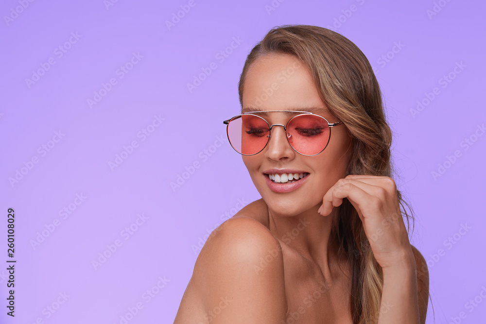 Portrait of beautiful young woman with perfect makeup wearing pink sunglasses. Smiling fashion model in aviator sunglasses posing on lilac background. Studio shot. Summer vacation.