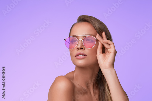 Portrait of beautiful young woman with perfect makeup wearing pink sunglasses. Smiling fashion model in aviator sunglasses posing on lilac background. Studio shot. Summer vacation.