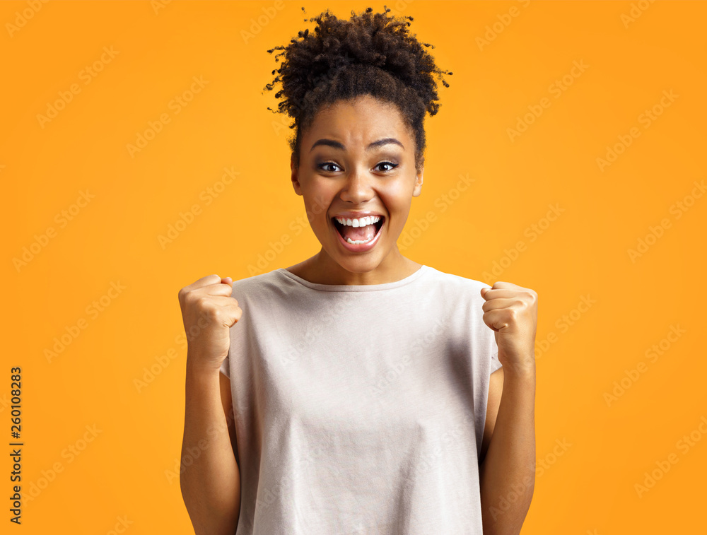 Overjoyed girl clenches fists with happiness, widely opens mouth as shouts loudly. Photo of african american girl wears casual outfit on orange background. Emotions and pleasant feelings concept. Overjoyed girl clenches fists with happiness, widely opens mouth as shouts loudly. Photo of african american girl wears casual outfit on orange background. Emotions and pleasant feelings concept.