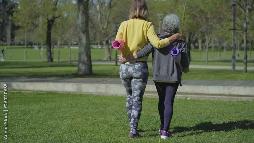 Rear view of young woman in yellow sweatshirt and middle-aged woman with grey hair walking in park, embracing each other, holding yoga mat in hands, talking. Sport, communication concept Rear view of young woman in yellow sweatshirt and middle-aged woman with grey hair walking in park, embracing each other, holding yoga mat in hands, talking. Sport, communication concept