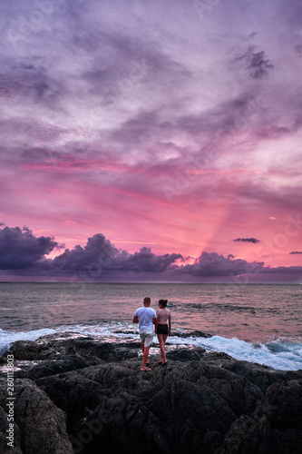 romantic couple standing on rocks by sea on purple dramatic sunset background