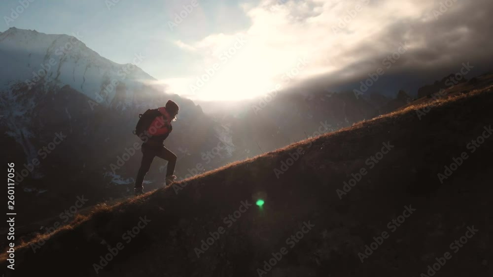Very long shot Aerial view of epic shot of a girl walking on the edge of the mountain as a silhouette in a beautiful sunset. Silhouette of a girl in a hat with a backpack climbing uphill