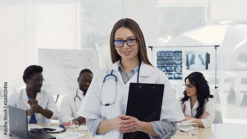 Young female doctor in white coat with stethoscope smiling looking into camera on colleagues background in clinic. Concept of doctors health care, love of medicine Young female doctor in white coat with stethoscope smiling looking into camera on colleagues background in clinic. Concept of doctors health care, love of medicine