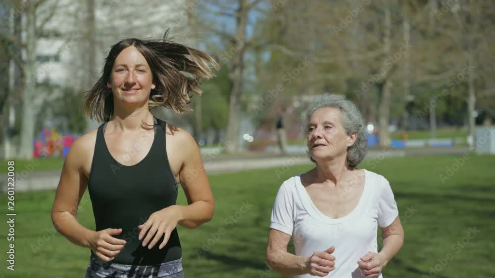 Front view of happy young woman in yellow sweatshirt and excited middle-aged woman with grey hair in white T-shirt jogging in park, talking, smiling. Medium shot. Sport concept Front view of happy young woman in yellow sweatshirt and excited middle-aged woman with grey hair in white T-shirt jogging in park, talking, smiling. Medium shot. Sport concept