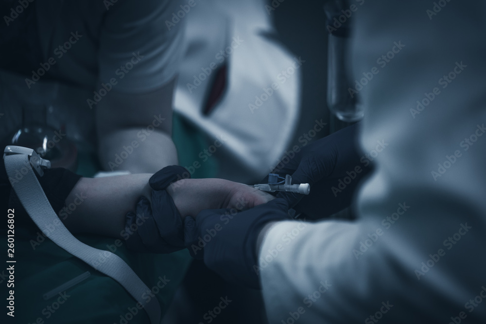 Close up picture of a young patient's hand while the doctors inject the anestetic agent before surgical intervention in a dark, creepy blue lighted surgery block