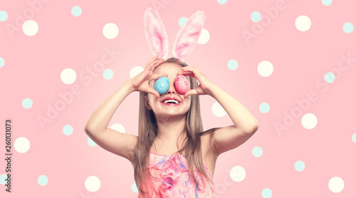 Funny Easter smiling little girl wearing bunny rabbit ears, holding colorful painted Easter eggs on her eyes. Baby girl laughing portrait over Polka Dots pink background