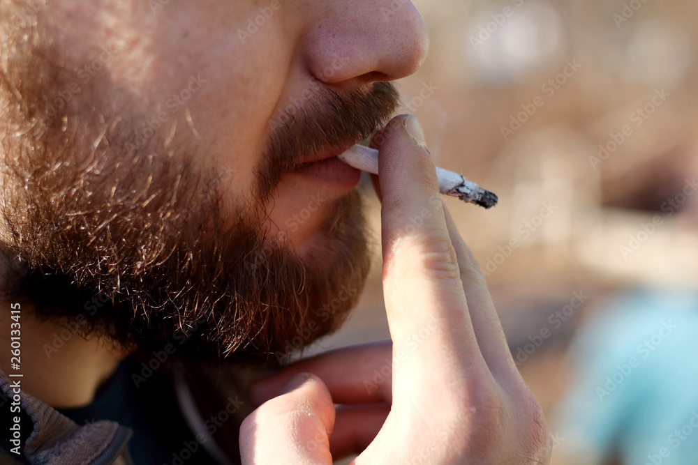 Fototapeta premium Portrait of brutal adult working man after labor day with a beard close up smoking cigarette, unheatlhy living