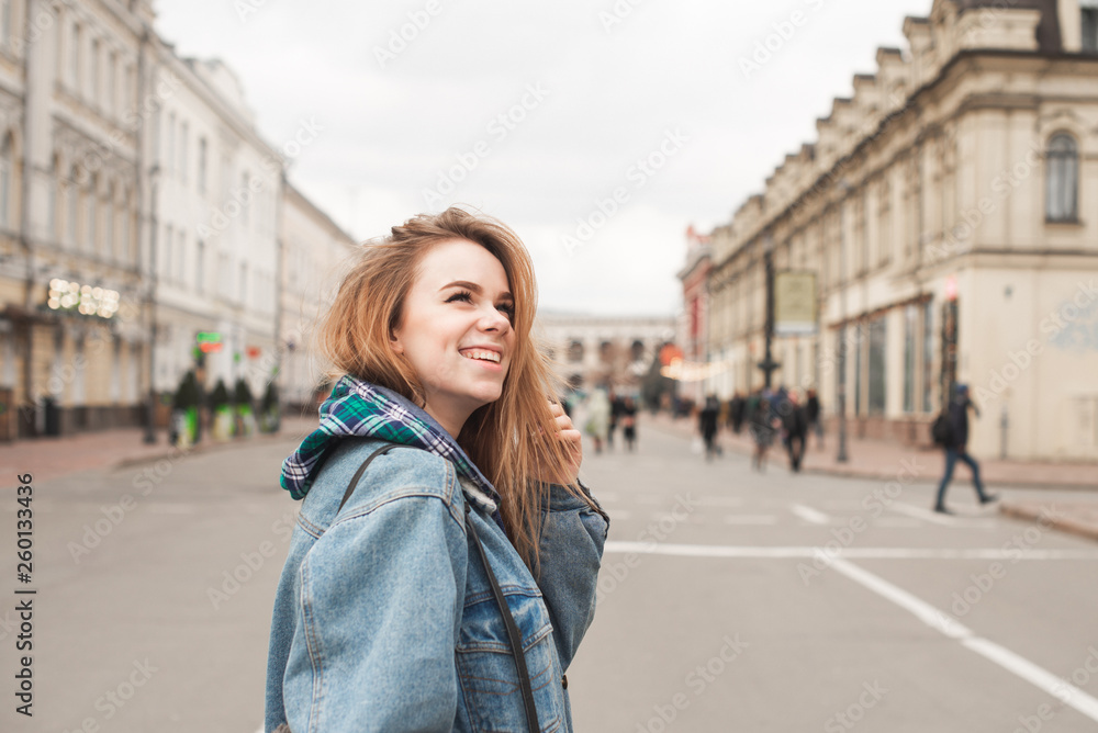 Smiling attractive girl walking along the street of the town, wearing a casual dress jeans jacket, looking sideways, background of the city landscape. Beautiful girl student on the streets of the city