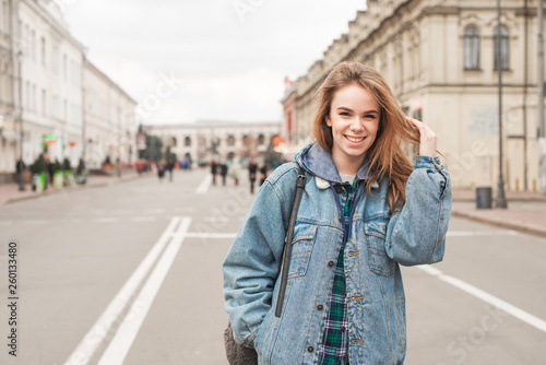 Happy girl in casual clothing is standing on the street of the town, smiling and looking at the camera. Positive student girl with a backpack against a street landscape. Street portrait. © bodnarphoto