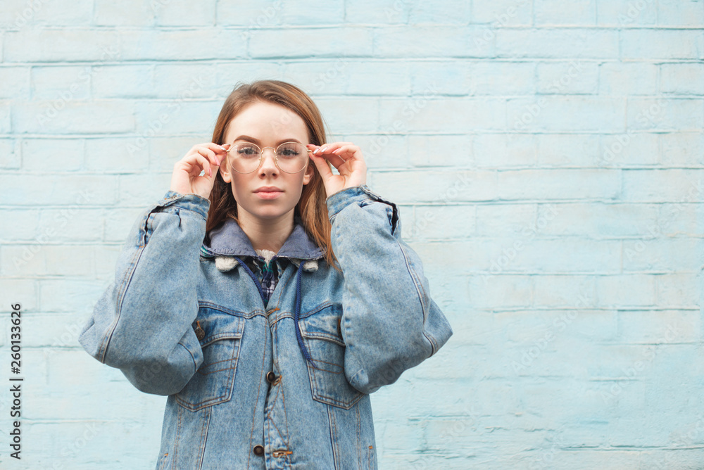 Obraz premium Beautiful girl in a denim jacket stands against the background of a blue wall, looks at the camera and fixes the glasses. Stylish hipster girl posing on a blue background. Copyspace