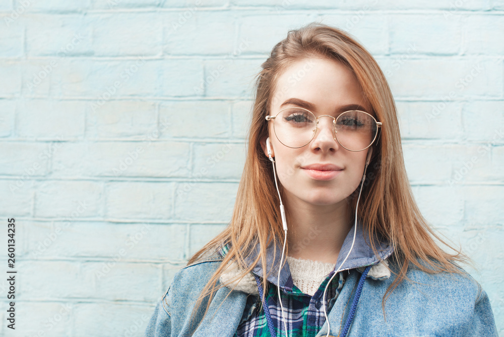 Beautiful girl in the headphones is standing against the background of a blue wall, looking into the camera with a serious face and listening to music. Close up portrait. Copyspace. Beautiful girl in the headphones is standing against the background of a blue wall, looking into the camera with a serious face and listening to music. Close up portrait. Copyspace.