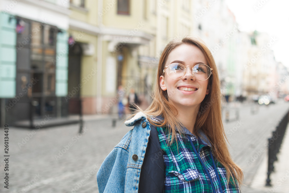 Happy student girl in casual clothing, wearing a denim jacket, glasses and a shirt, standing on the back street of the town, looking sideways and smiling. Portrait of a teenage tourist.