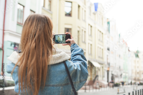 Back of a stylish girl makes a photo of the street of a European town on a smartphone. Tourist girl photographs a city landscape on a smartphone. Focus on a smartphone with a camera.
