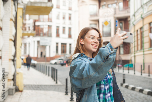 Happy girl traveler in casual clothes walks in the old town and takes a photo landscape on a smartphone. Beautiful tourist in a jeans jacket photographs a city landscape on the phone and smiles.