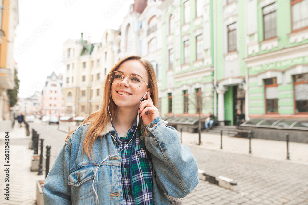 Fototapeta premium Stylish cheerful girl walking along the streets of the town with music in the headphones, smiling and looking away, wearing a jeans jacket. Street student portrait of a student with headphones