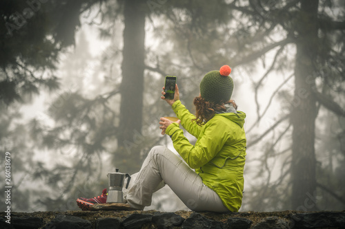 Mountain trekking lover woman makes a break drinking hot coffee and making a video call with remote friends. Sporty girl sitting on a wall in a misty forest shooting selfies. Winter autumn landscape