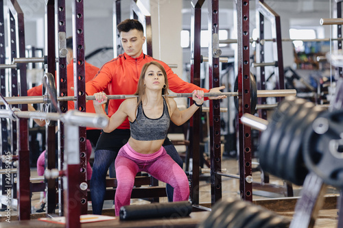 young girl doing squats with a barbell in the gym under the supervision of a trainer