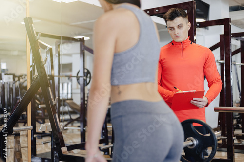 young girl doing squats with a barbell in the gym under the supervision of a trainer