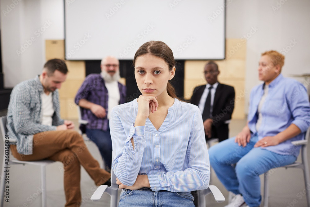 Portrait of pale young woman looking at camera while sitting in chair in front of therapy group, copy space Portrait of pale young woman looking at camera while sitting in chair in front of therapy group, copy space