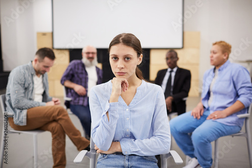 Portrait of pale young woman looking at camera while sitting in chair in front of therapy group  copy space