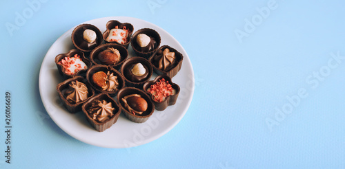 Beautiful chocolates of different shapes and fillings lie in a white plate against the blue background paper. Close up, copy space.