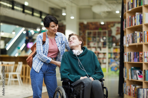 Foto Positive attractive young woman with satchel pushing wheelchair and talking to d