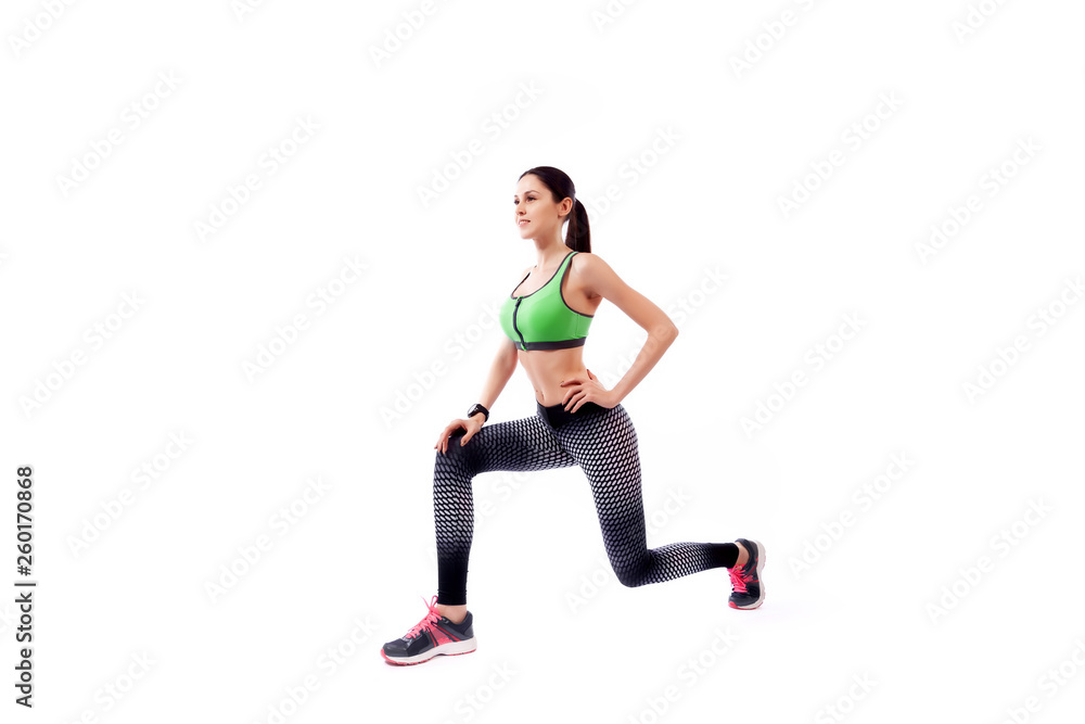 A young woman model in a sporty short top and gym leggings makes lunges by the feet forward, hands are held out to the side on a white isolated background in studio A young woman model in a sporty short top and gym leggings makes lunges by the feet forward, hands are held out to the side on a white isolated background in studio