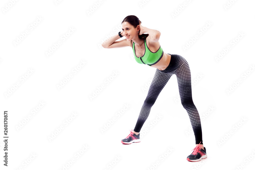 A young woman model in a sporty short top and gym leggings makes bends forward, arms behind head, legs wide apart  on a  white isolated background in studio