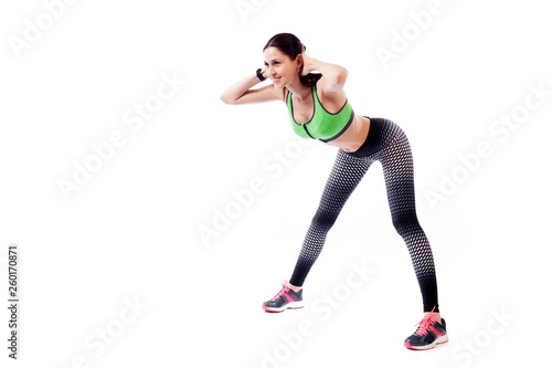 A young woman model in a sporty short top and gym leggings makes bends forward, arms behind head, legs wide apart on a white isolated background in studio